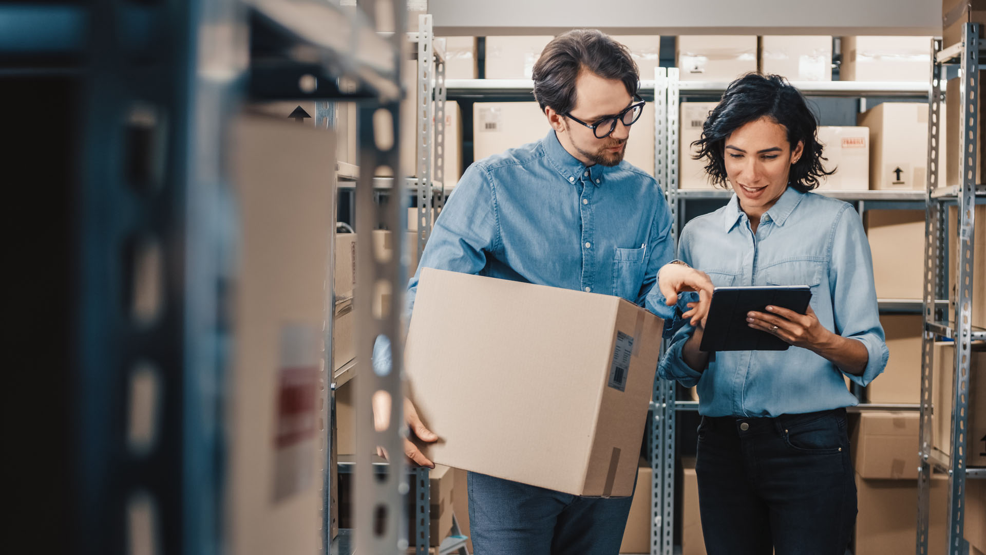 Female Inventory Manager Shows Digital Tablet Information to a Worker Holding Cardboard Box, They Talk and Do Work. In the Background Stock of Parcels with Products Ready for Shipment.